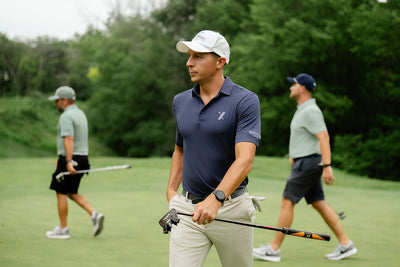 Man in a blue shirt and white cap walking on a golf course with other golfers in the background.