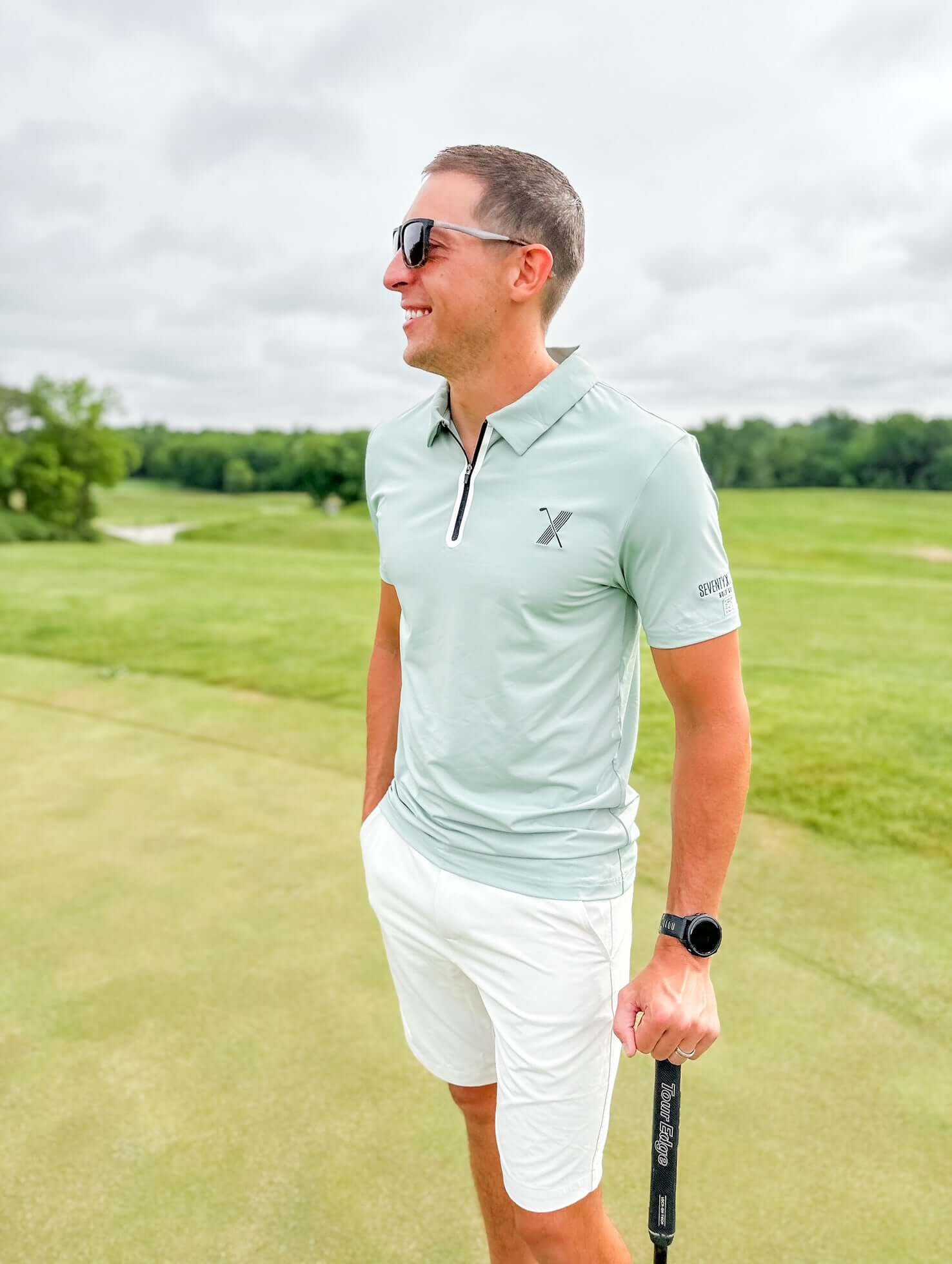 Man wearing light green Forgiveness Polo and black hat sitting at golf course table, smiling