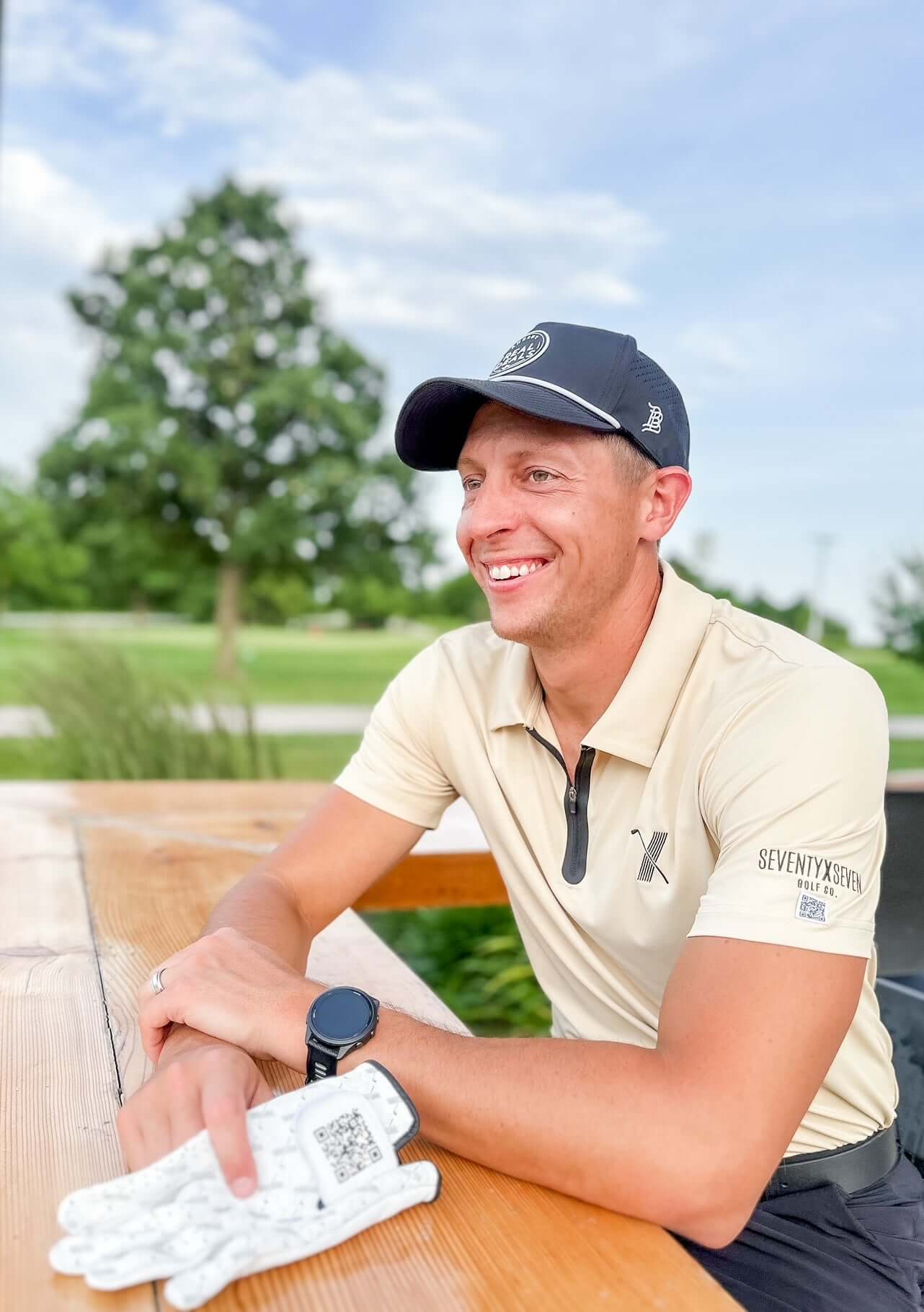 Man wearing cream Forgiveness Polo and black hat sitting at golf course table, smiling